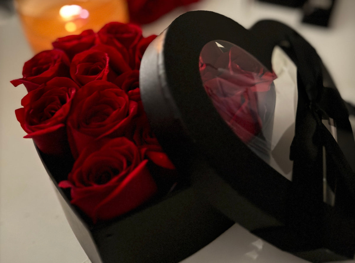A heart-shaped black gift box filled with deep red roses, with the lid propped open to reveal a clear window showing a wrapped red rose inside. A warm blurred candle glows in the background.