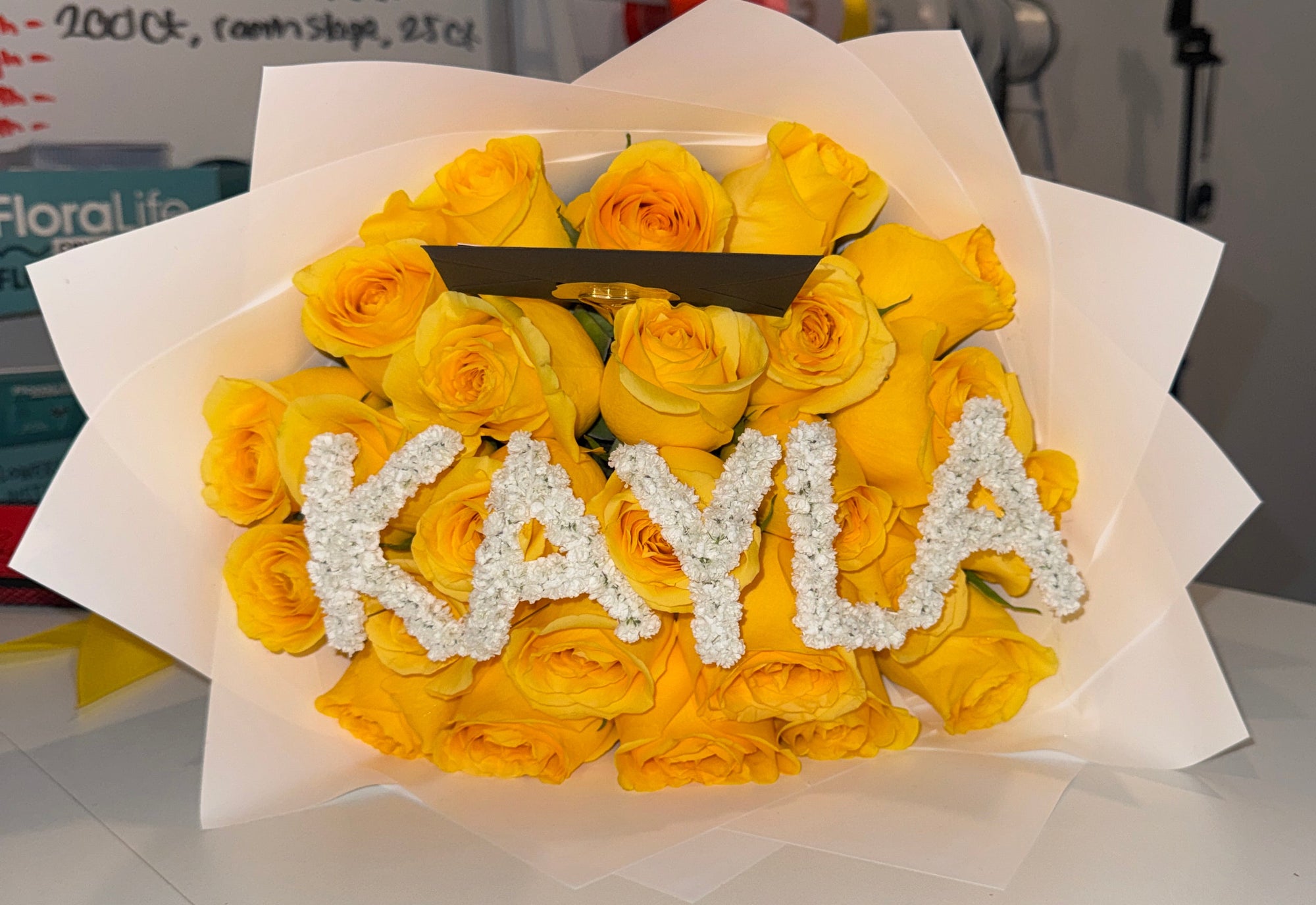 A bouquet of yellow roses wrapped in white tissue paper, with the name 'KAYLA' spelled out in white rhinestone letters across the front. The bouquet sits on a white table in an indoor setting.
